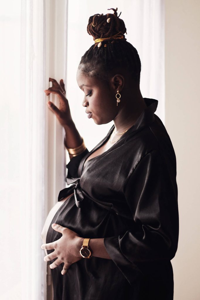 Séance photo de femme enceinte à domicile regardant vers son ventre, devant la fenêtre