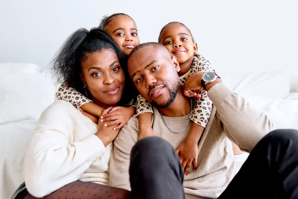 Dawakeen-photographe-yvelines-portrait-famille 3 Portraits de famille noire à domicile, les parents assis au sol et les filles derrière les parents leur faisant un câlin à chacun