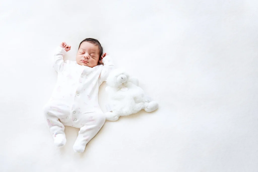 Photographie d'un nouveau-né allongé sur le dos, les bras en l'air, à côté de son doudou dans la même position