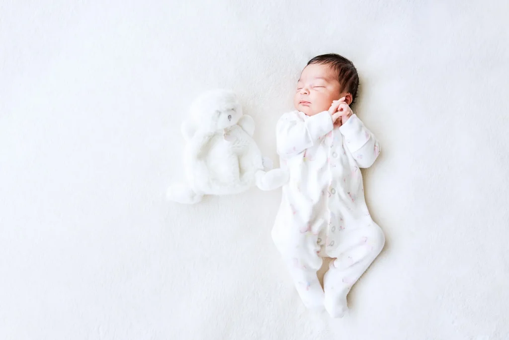 Photographie d'un nouveau-né allongé sur le dos, les mains sur le côté de sa joue et un doudou à côté
